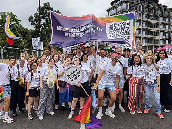 Group of researchers attending the Pride in London march in London, July 2025, wearing white T-shirts printed with the MRS logo in rainbow colours and holding Market Research Society sign, a banner and rainbow flags.